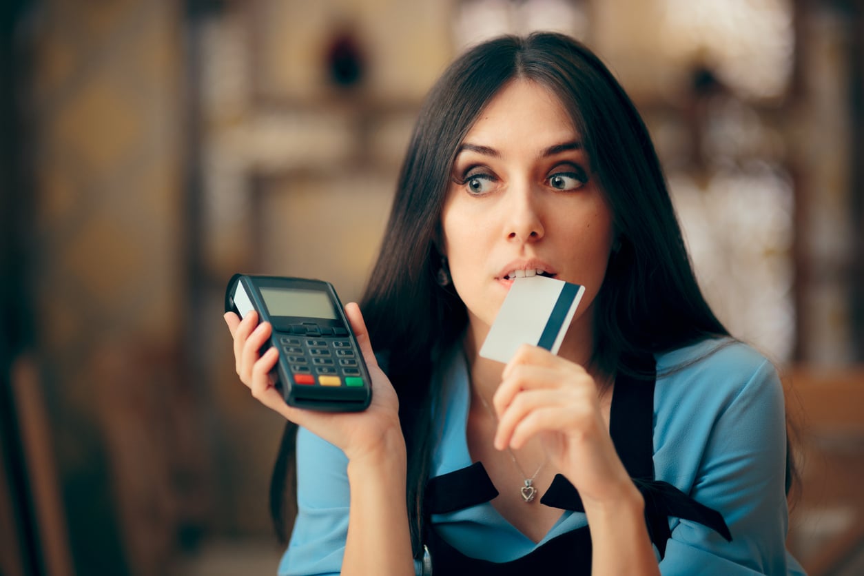 A woman wearing an apron holds a credit card terminal in one hand and bites the edge of a credit card with a worried expression.