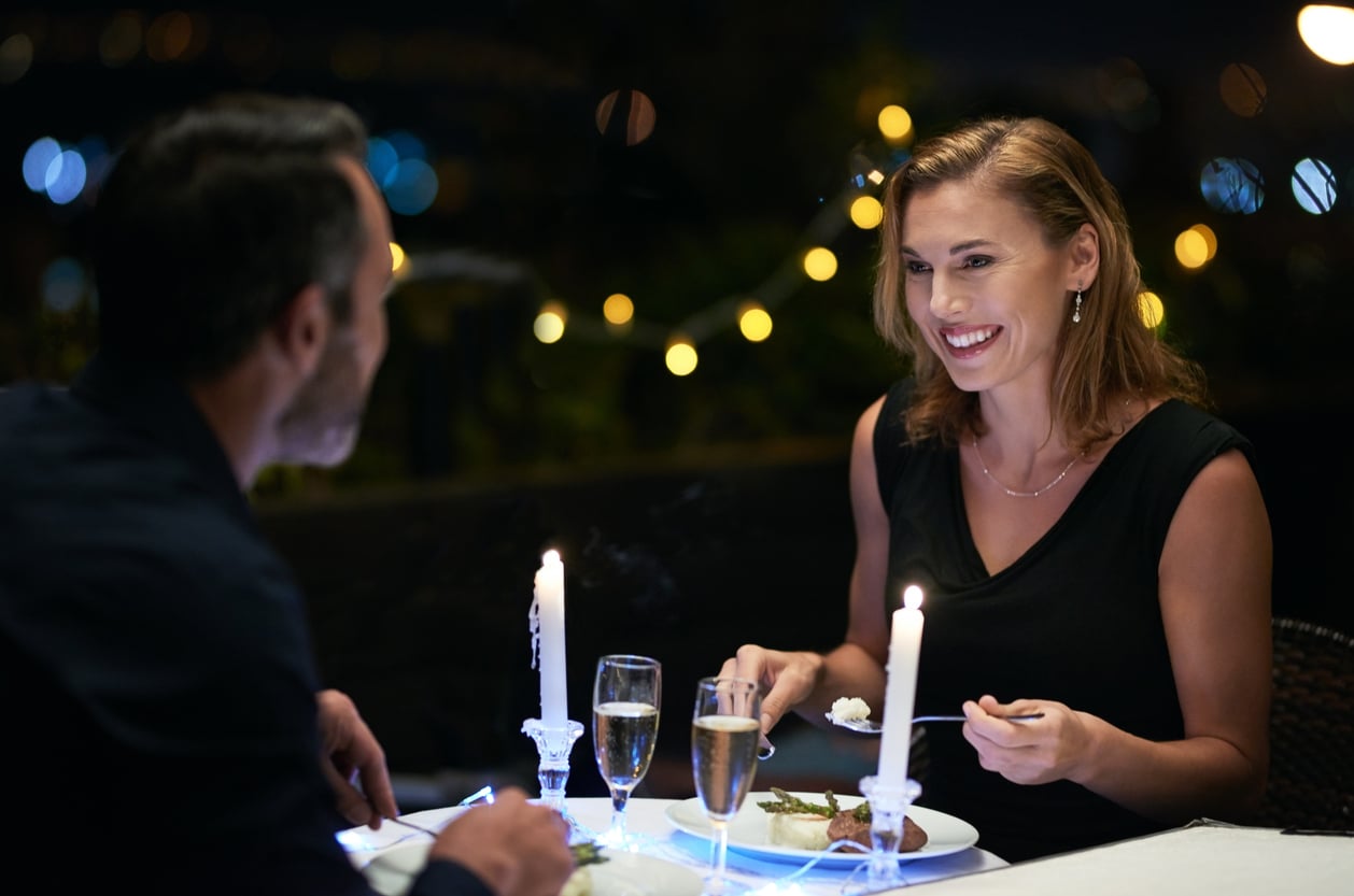 A smiling woman and a man enjoy a candlelit dinner outdoors at night.