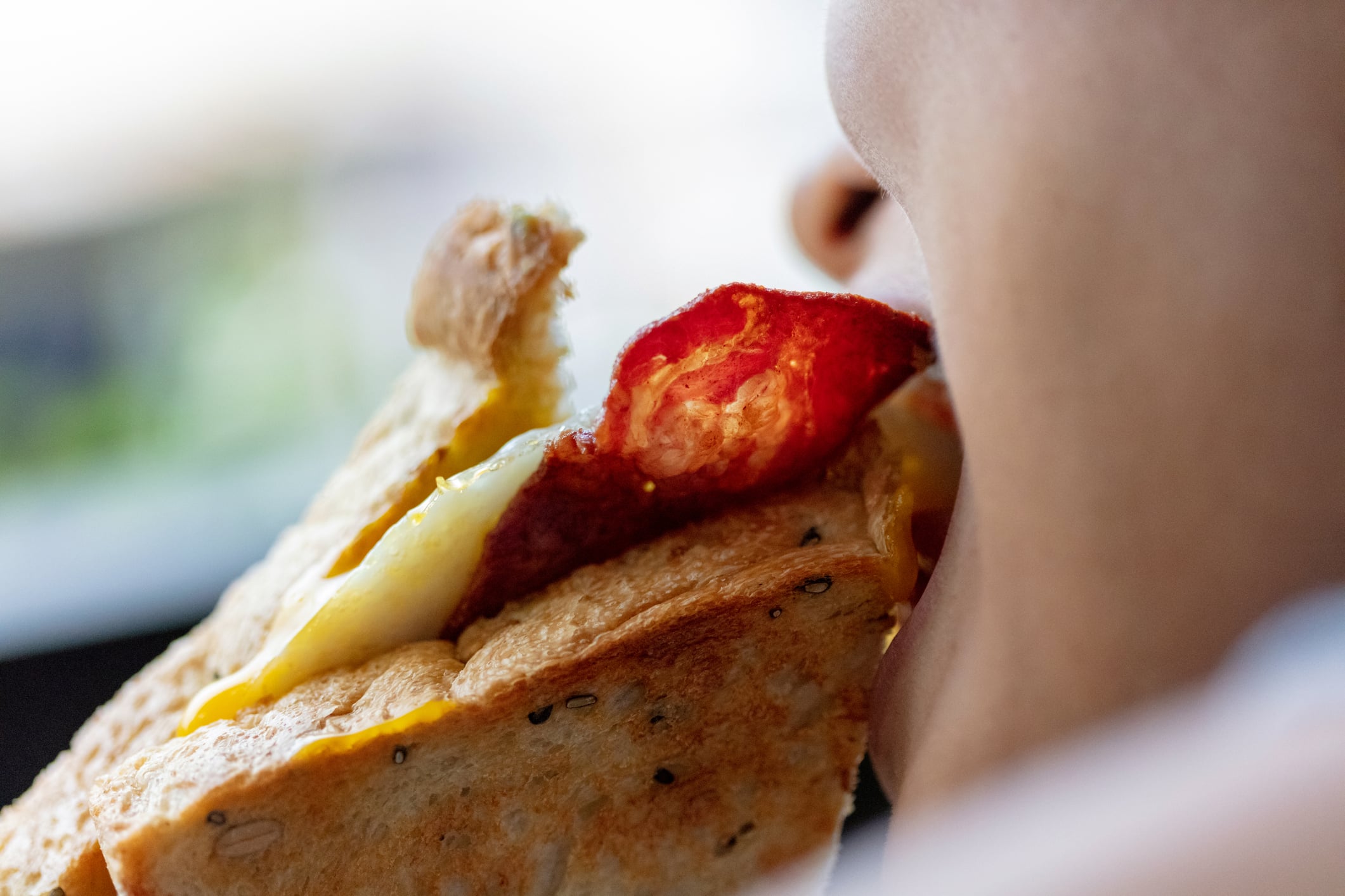 A person taking a bite of a toasted egg and cured meat breakfast sandwich.