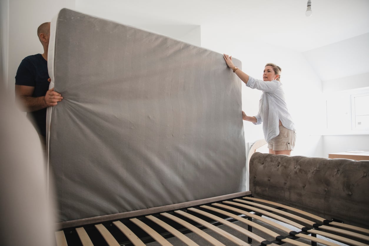 A man and a woman stand on opposite sides of a bed frame, lifting and positioning a large mattress onto the slats.