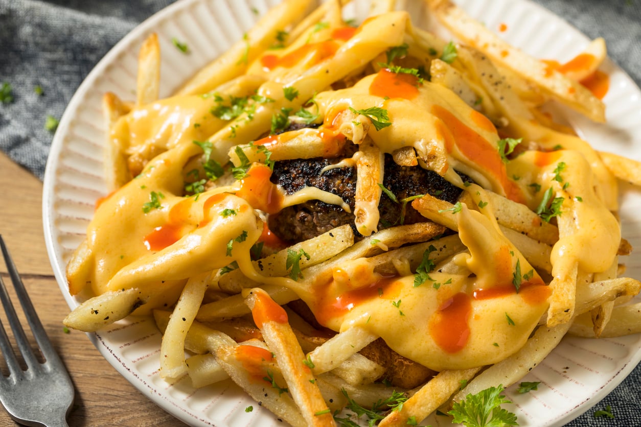 A plate of french fries topped with melted cheese, hot sauce, and herbs, covering a hamburger patty.
