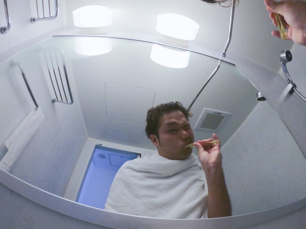 A man wearing a towel around his shoulders brushes his teeth while looking into a bathroom mirror.