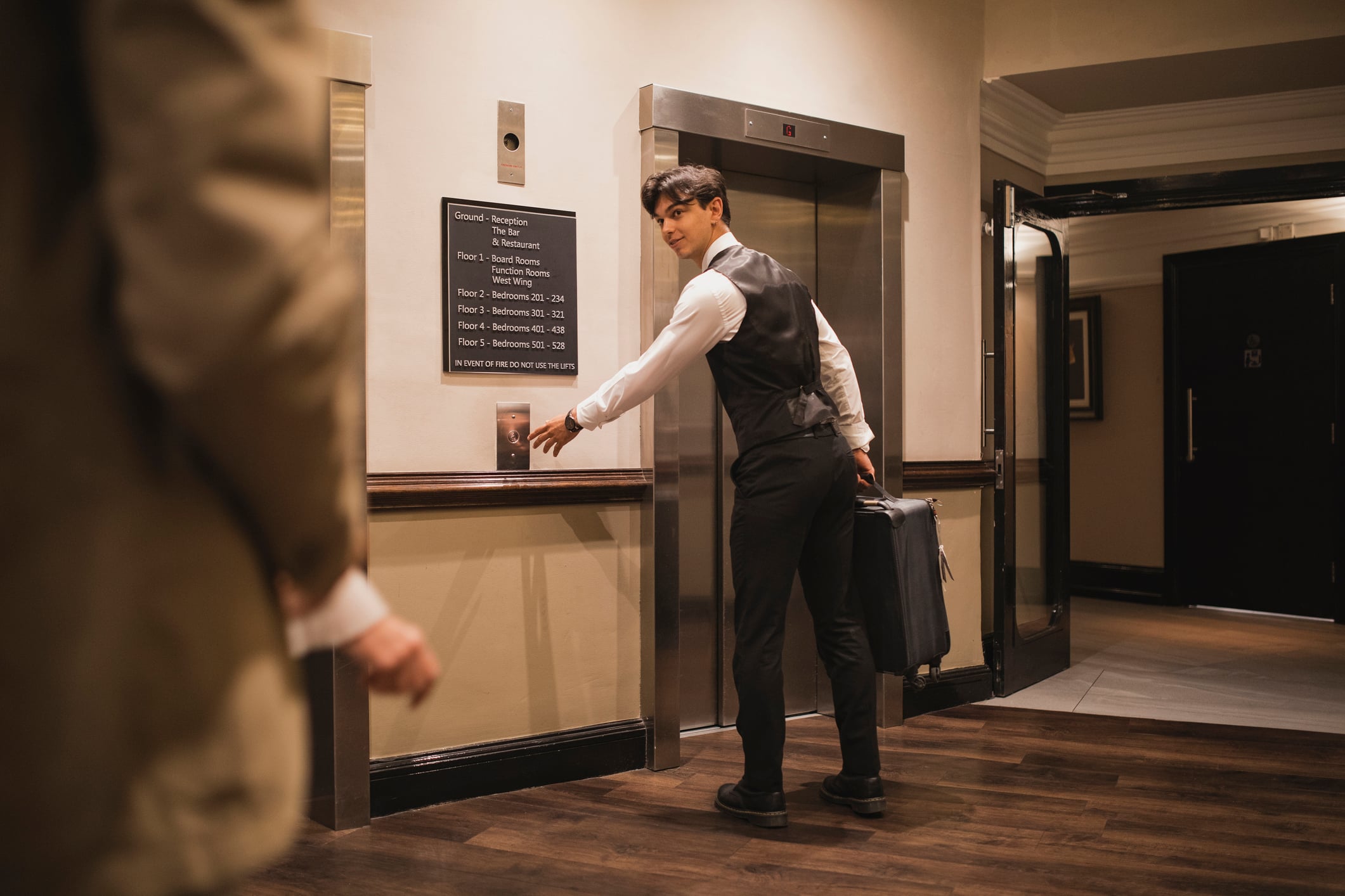 A bellhop in a vest and dress shirt presses the button for an elevator in a hotel hallway, in one of the hotel traditions.