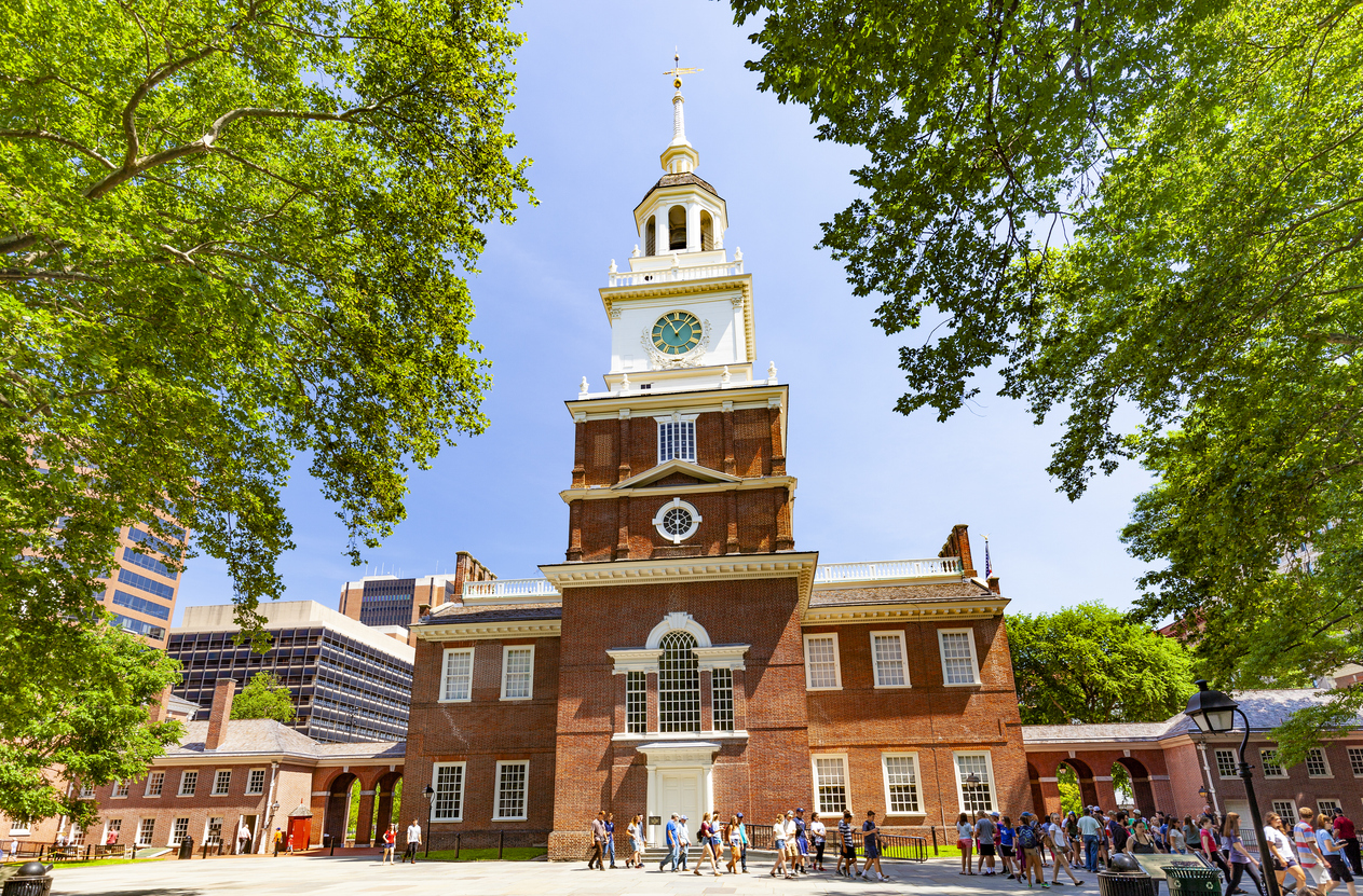 Independence Hall in Philadelphia, a historic red-brick building with a clock tower, surrounded by green trees and people gathered outside on a sunny day.