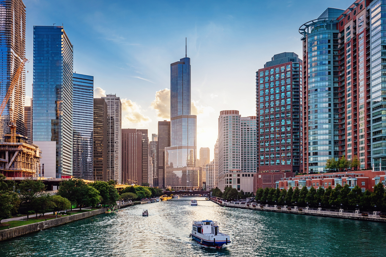 Boats travel along a river flanked by tall skyscrapers and modern buildings in a city at sunset, with the sky partly cloudy and sunlight reflecting off the glass structures.