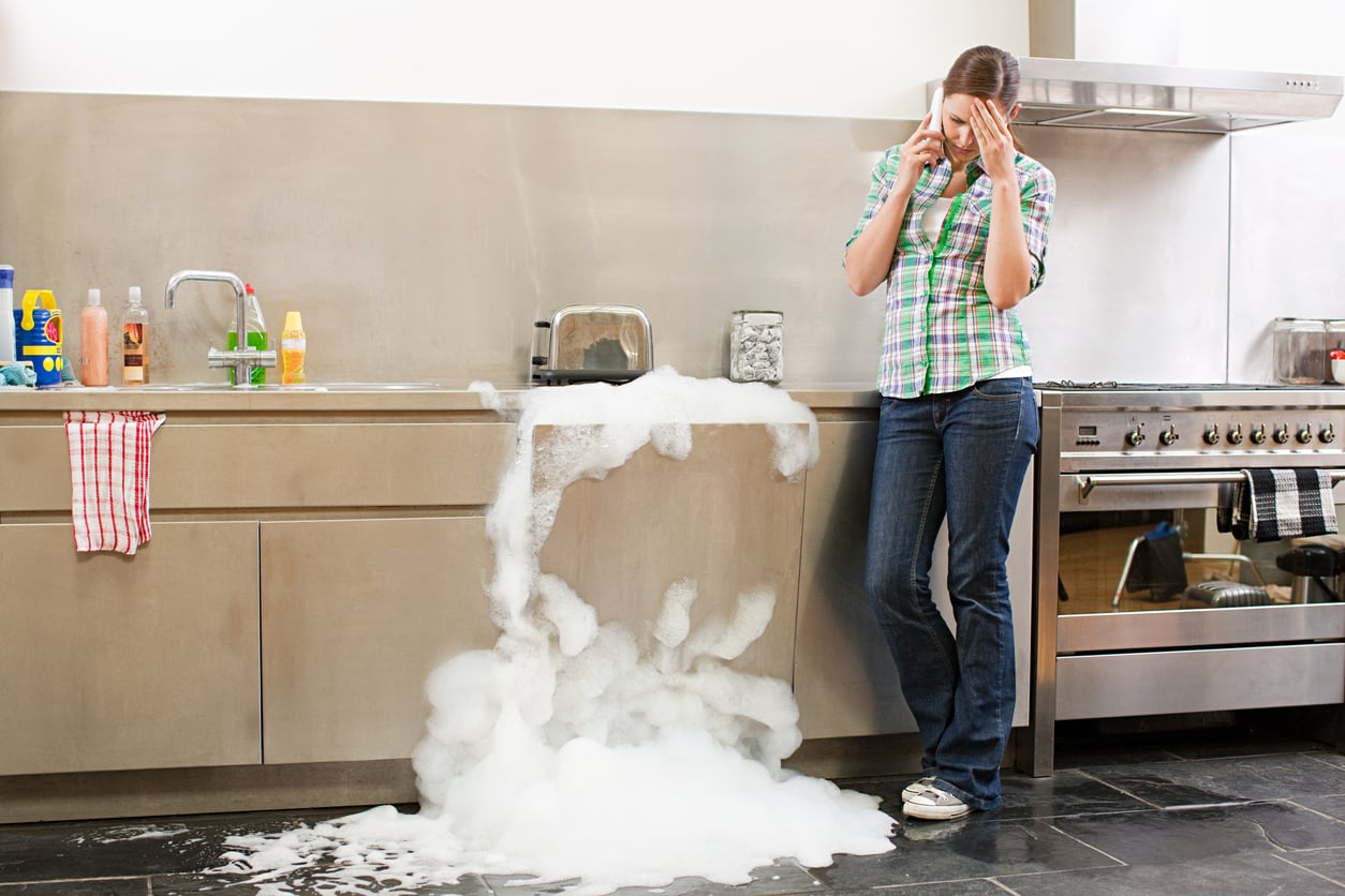 A woman leans against a kitchen counter looking stressed while talking on the phone, as thick soap suds overflow from the dishwasher onto the floor.
