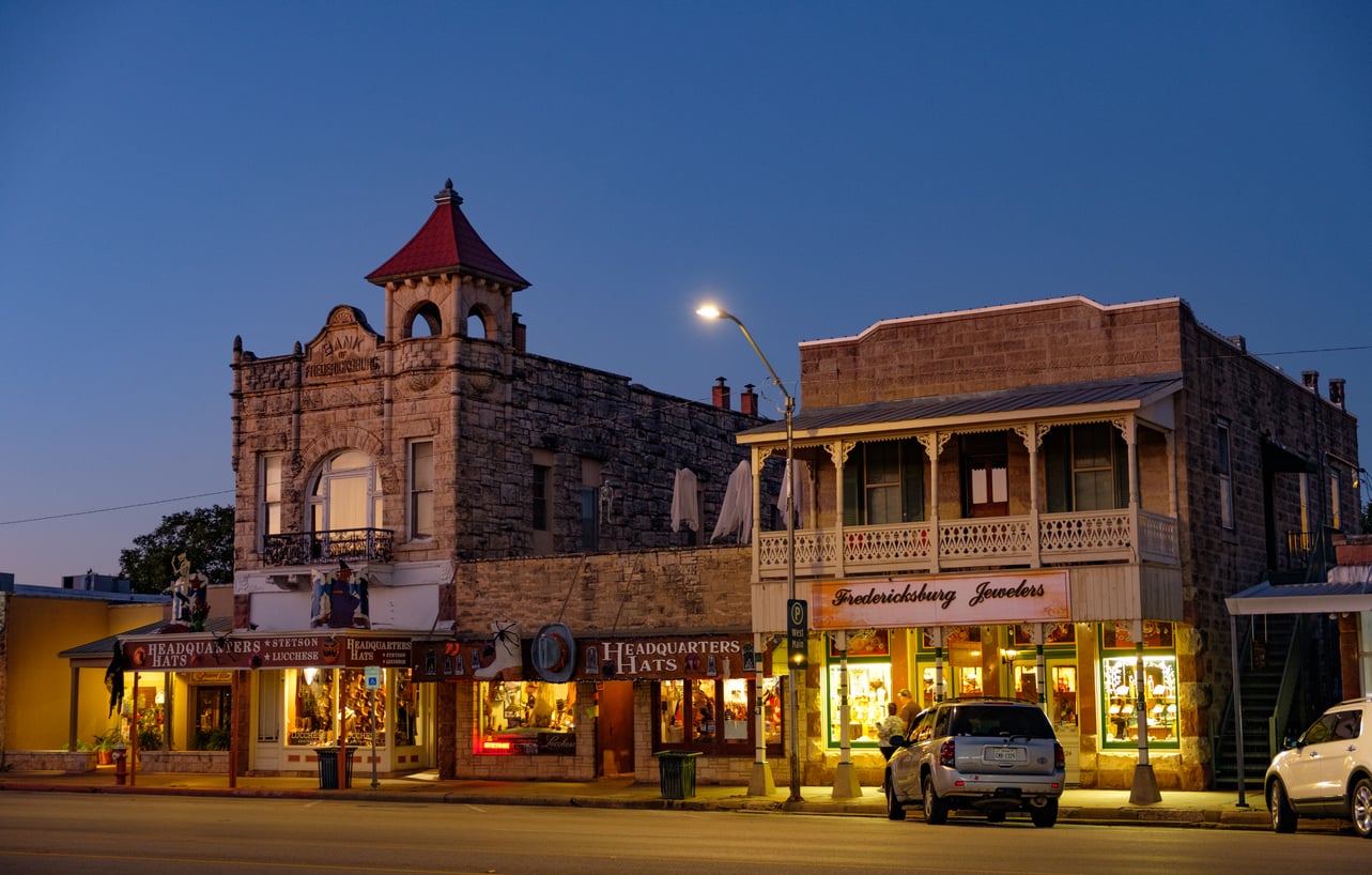 Two historic stone buildings with shops, hats, and jewelry stores lit up at dusk. A parked car sits outside, and a streetlamp glows as the sky darkens—one of those American Destinations That Feel Bigger.
