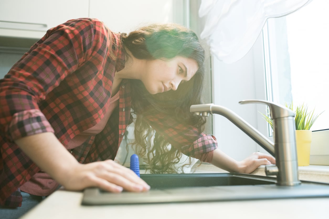 A woman leaning over a kitchen sink, looking down into the drain.