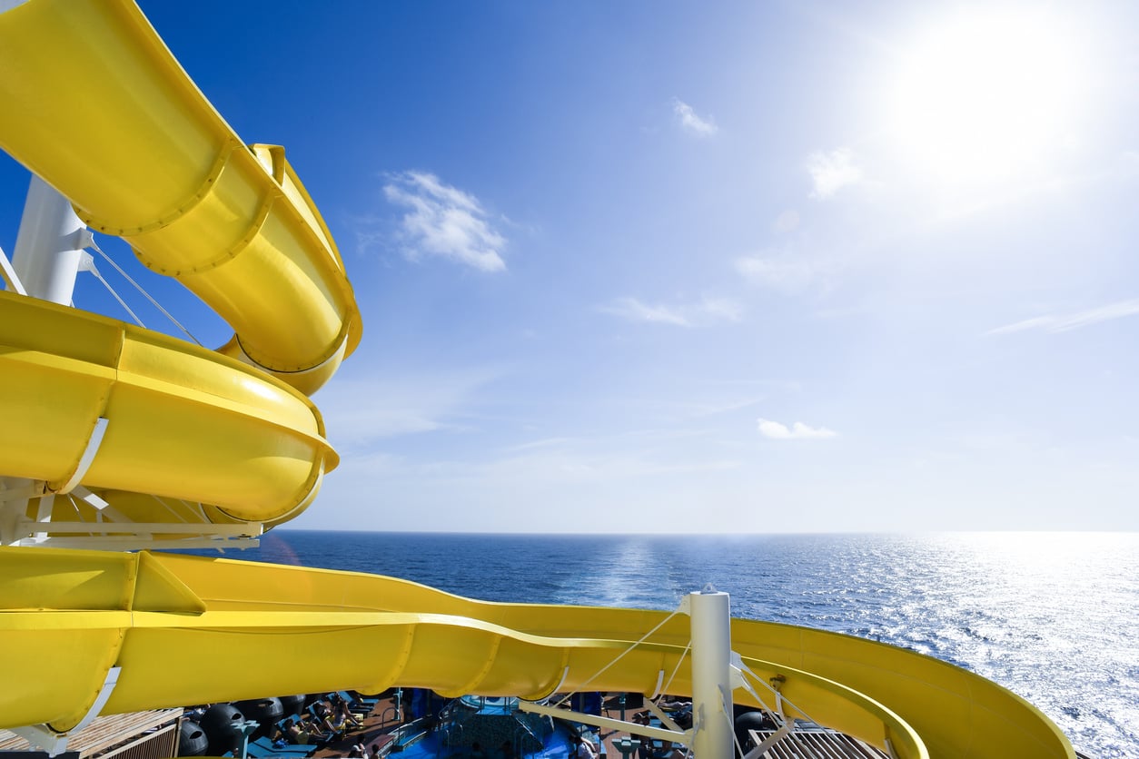 A bright yellow water slide curves high above the deck of a cruise ship, overlooking the ocean under a clear sky as one of many cruise ship amenities.