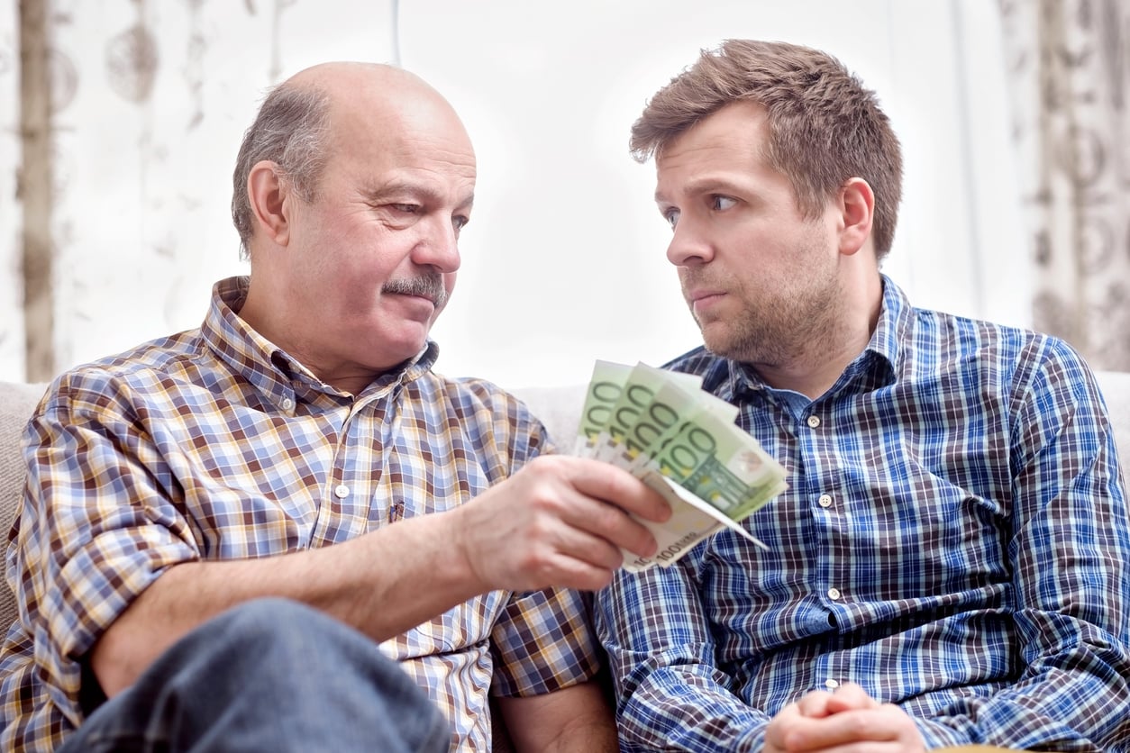 An older man in a plaid shirt hands financial help, a fan of 100 euro banknotes to a younger man.