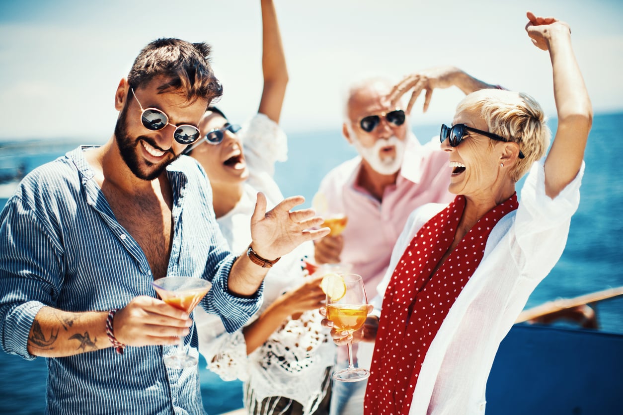 Four people in sunglasses dance and hold cocktails while on a boat at sea.