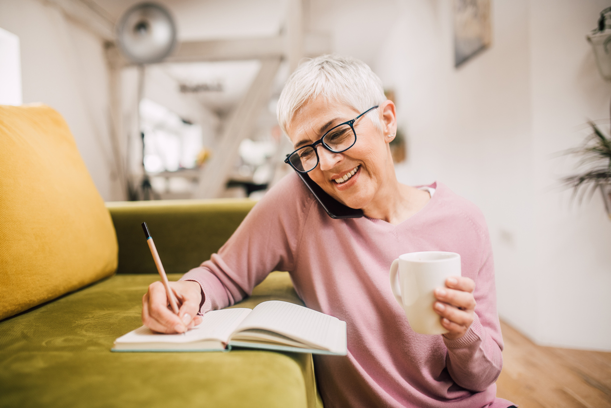 A woman with short gray hair wearing glasses talks on her phone while writing in a notebook and holding a mug.