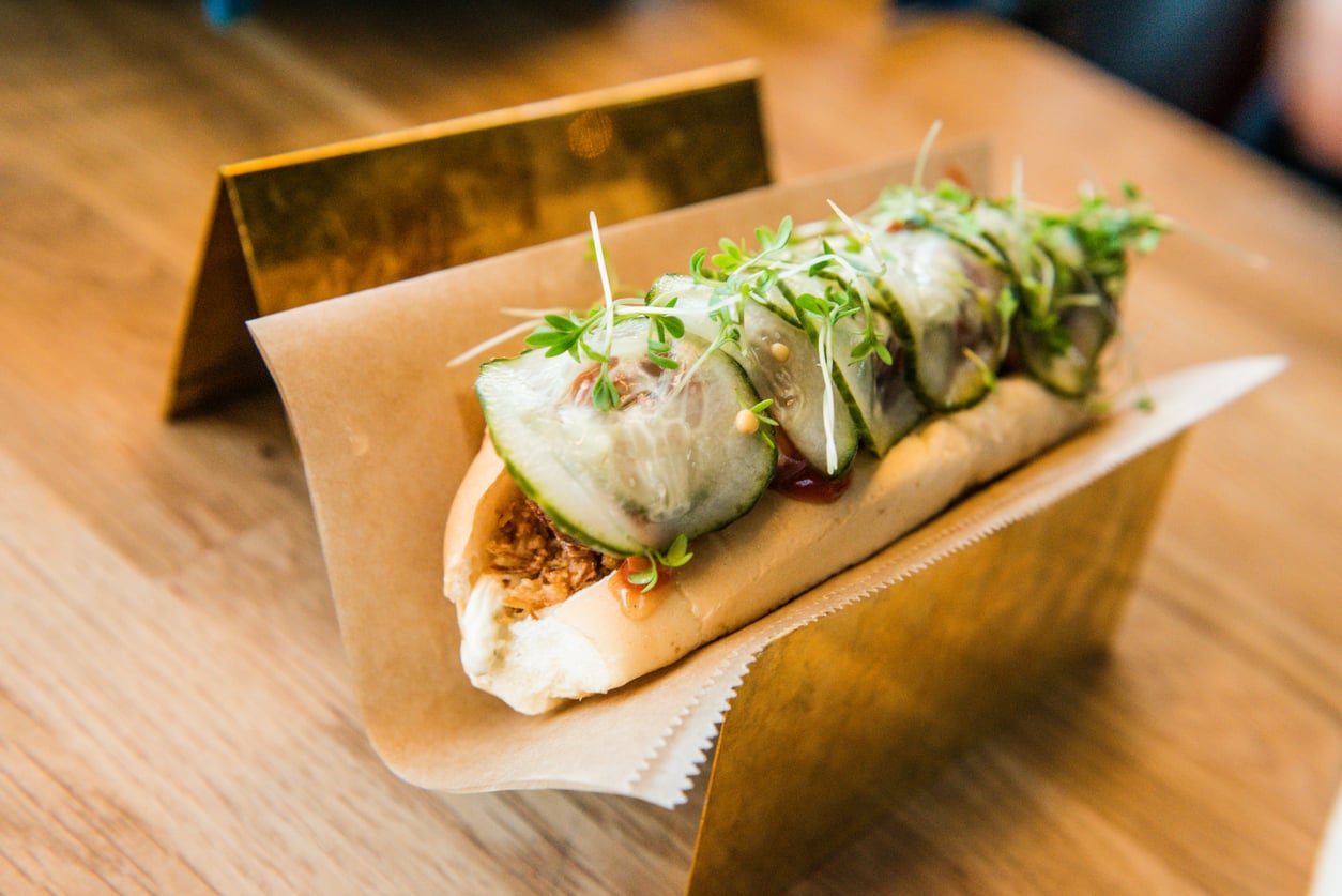 A hot dog in a toasted bun topped with thin cucumber slices and microgreens, served in a gold-colored metal stand.