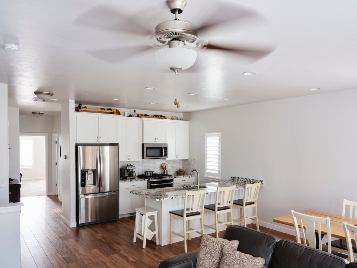 A kitchen with white cabinets, stainless steel appliances, a granite-topped island with three bar stools, and a ceiling fan.