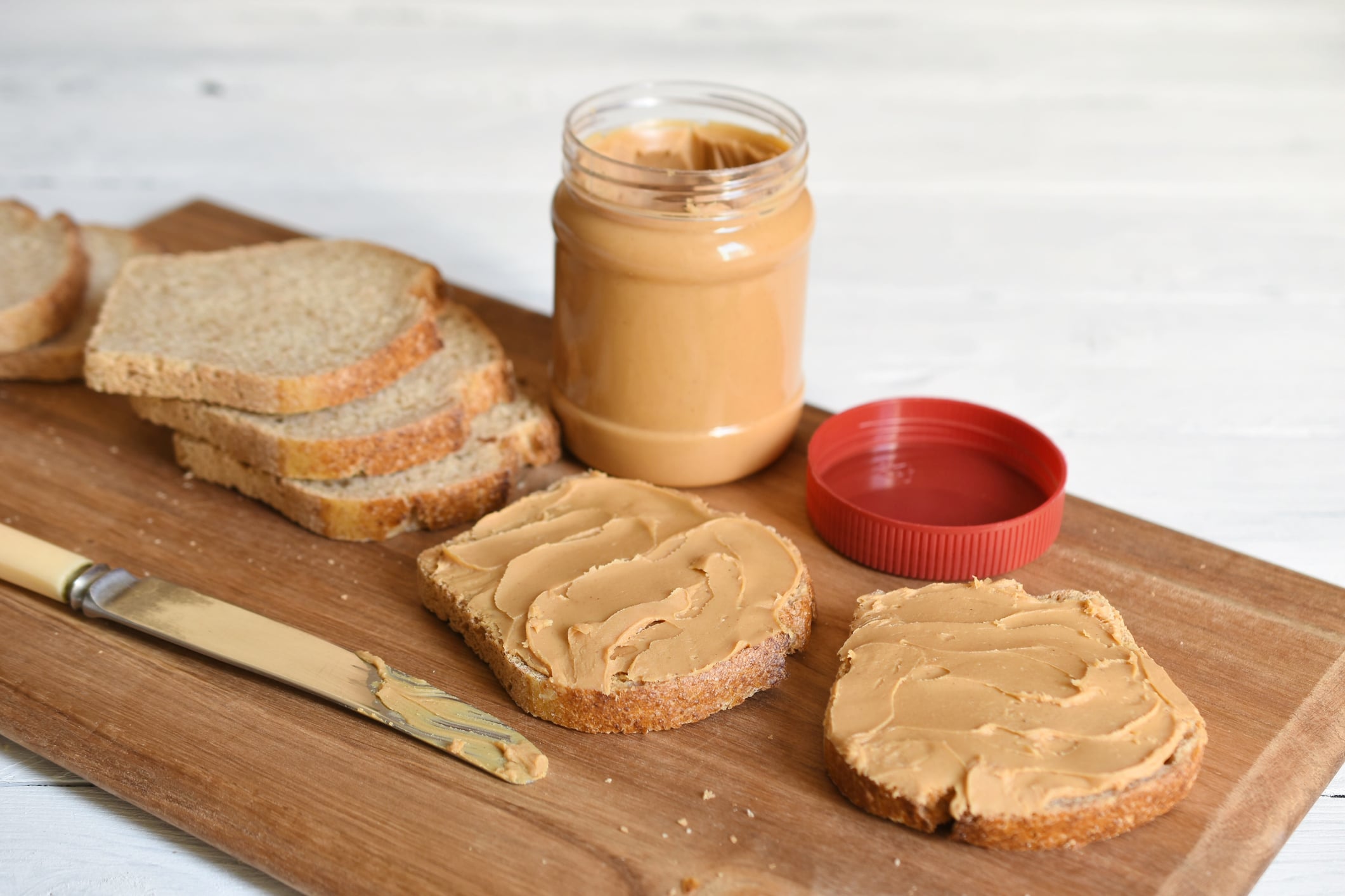 Two slices of bread spread with peanut butter, a jar of peanut butter with its lid nearby, and a butter knife on a wooden cutting board.
