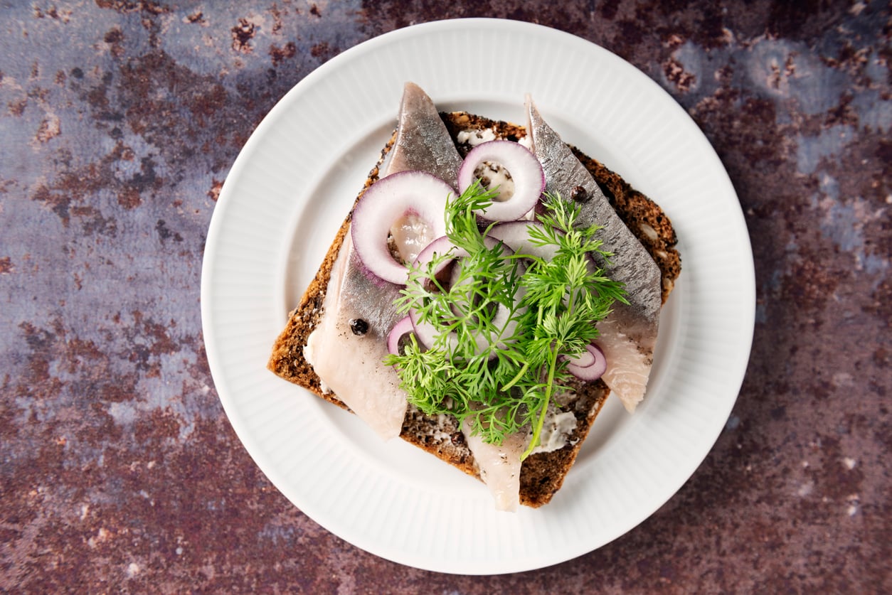 A slice of dark rye bread topped with herring, red onion rings, and fresh dill on a white plate.