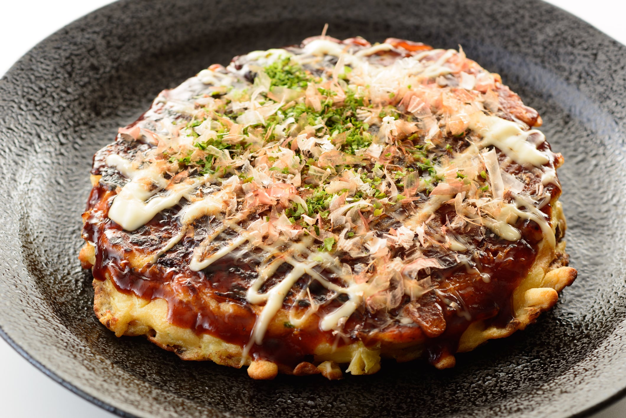 Okonomiyaki pancake topped with sauce, mayonnaise, dried seaweed, and bonito flakes on a dark textured plate.
