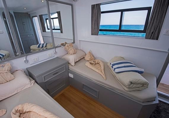 Interior of a boat cabin with two single beds, each featuring towel art, and a view of the ocean through a rectangular window.