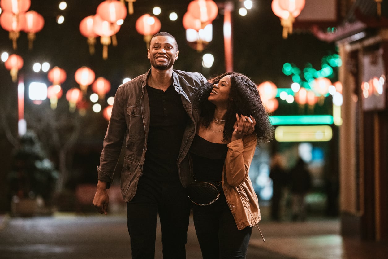 A smiling couple walks arm-in-arm down a street decorated with glowing red lanterns at night.