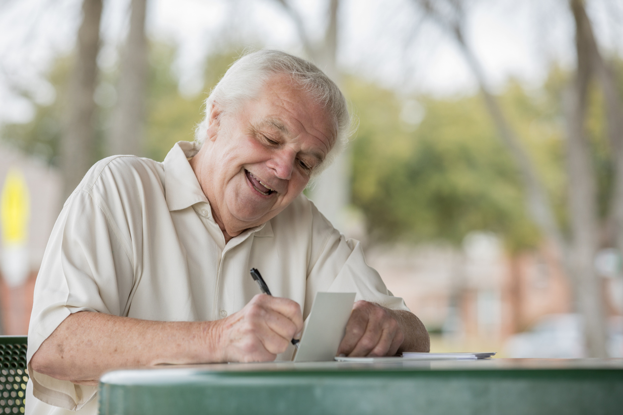 An older man in a light-colored shirt smiles while writing on a small card at an outdoor table.