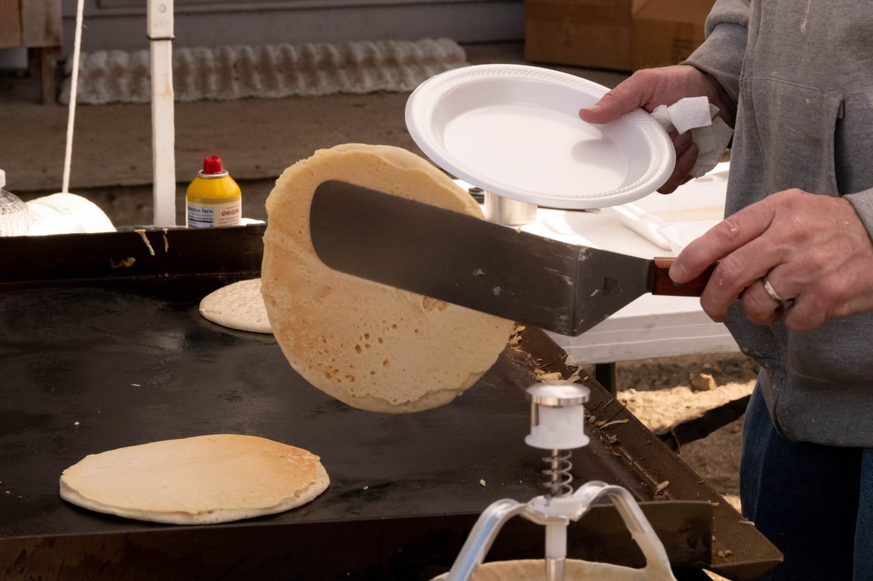 A person uses a metal spatula to flip a pancake on a flat-top grill while holding a white plate.