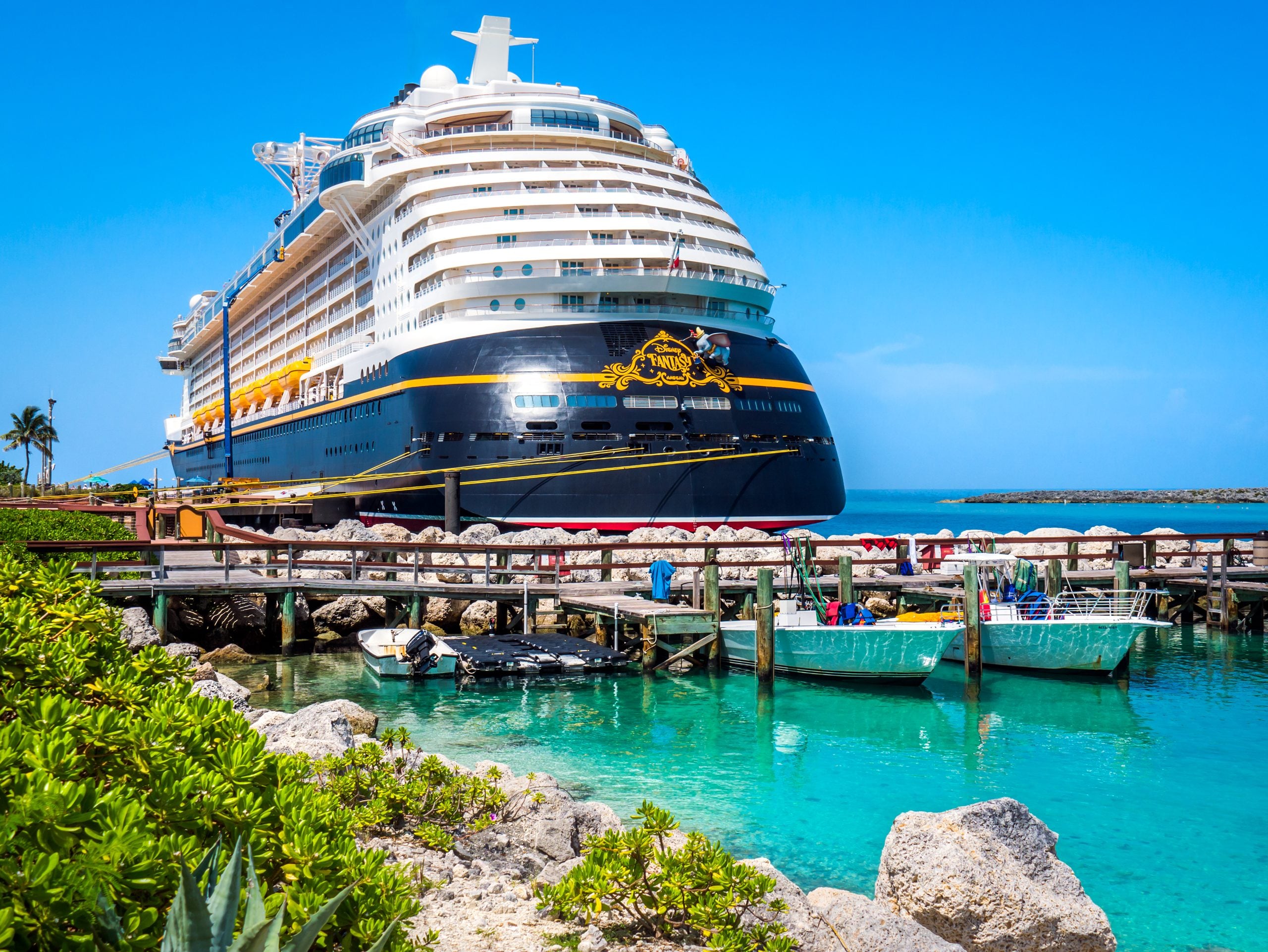 Disney Fantasy cruise ship docked at a tropical pier next to several small boats in turquoise water.