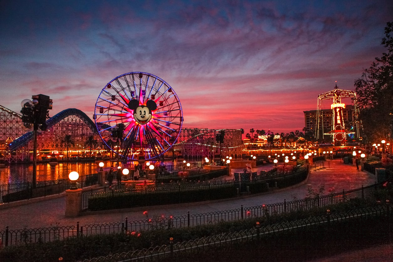 Pixar Pal-A-Round Ferris wheel with a Mickey Mouse icon at dusk, featuring a silhouetted amusement park and roller coaster against a vibrant pink and purple sky.