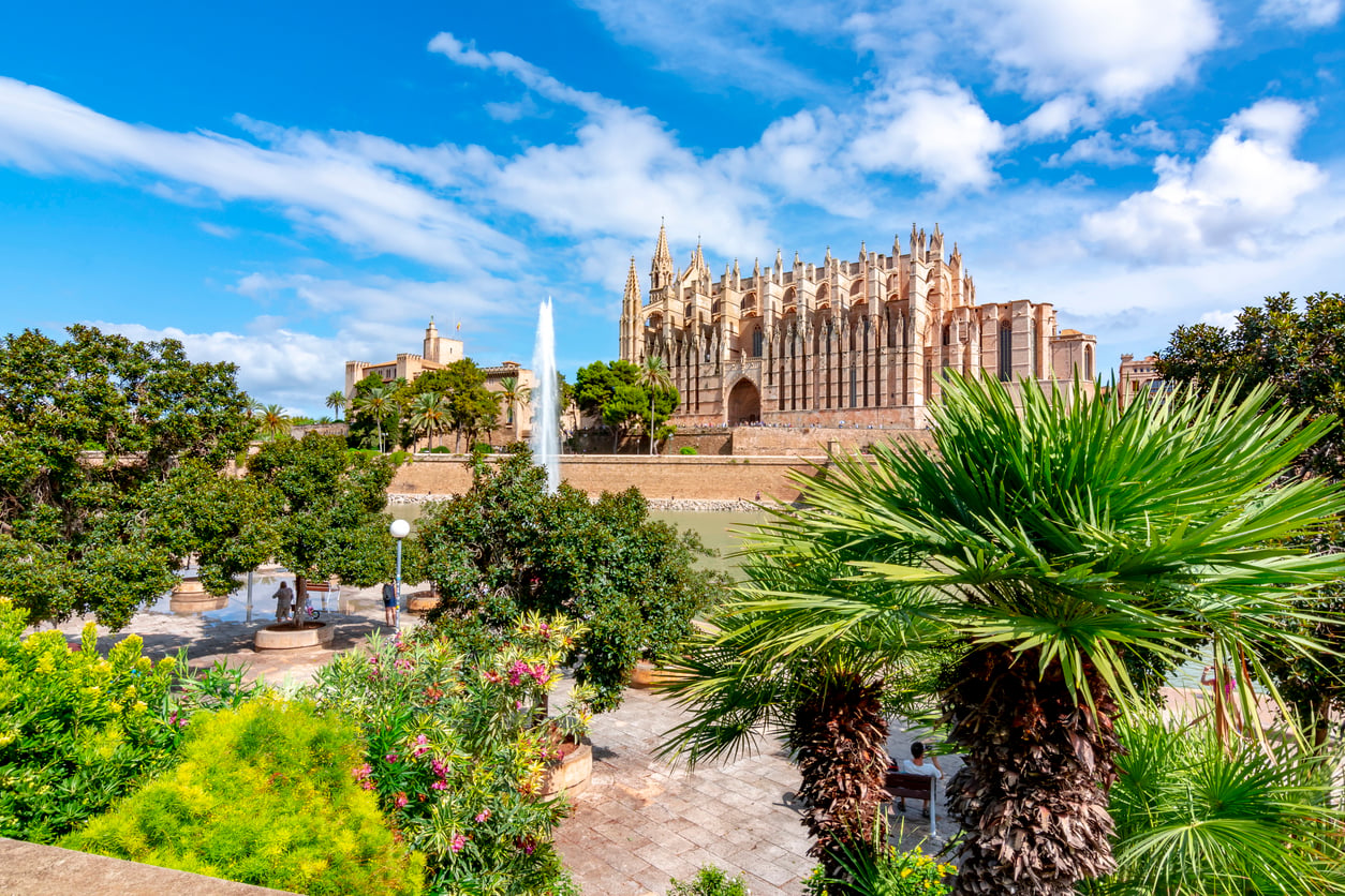 The Palma Cathedral stands prominently above a park featuring a fountain and lush trees under a bright blue sky.