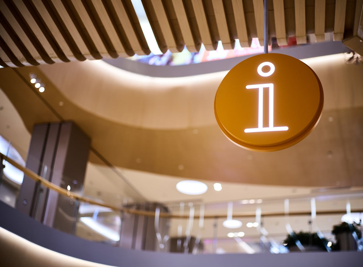 A circular, glowing yellow information sign suspended from the ceiling in a multi-level shopping center.