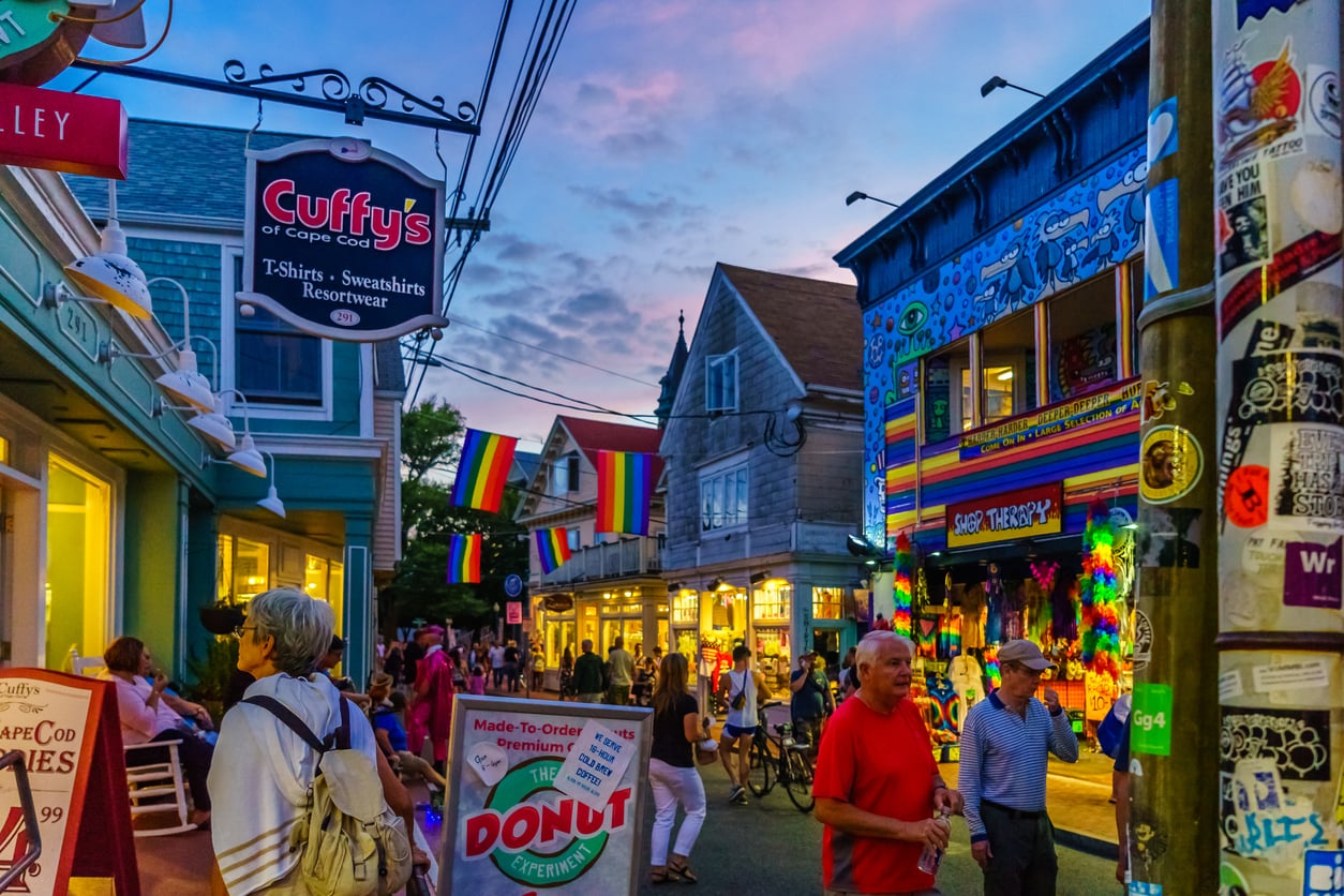 Colorful street scene at sunset with people walking, rainbow flags hanging, shops, and vibrant decorations. A donut sign and shop signs are visible, with lively, festive atmosphere and stickers on a nearby pole.