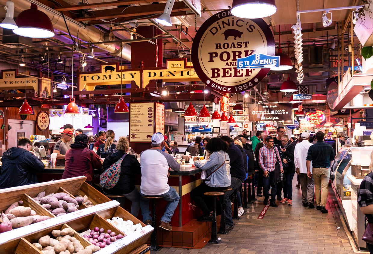 Customers at a crowded indoor public markets sitting at a counter labeled “Tommy DiNic’s,” with bins of potatoes in the foreground and a hanging sign for DiNic’s Roast Pork and Beef.