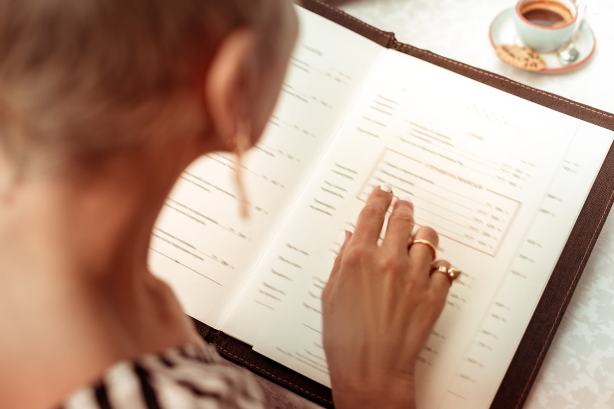 A person pointing to an item on a restaurant menu, with a small cup of espresso nearby.