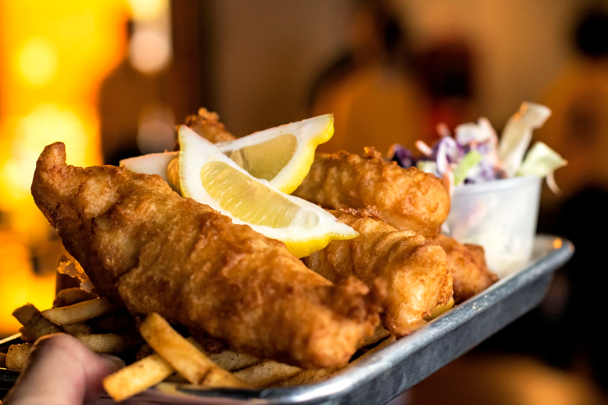 A metal tray of fish and chips topped with lemon wedges, accompanied by a side of coleslaw.