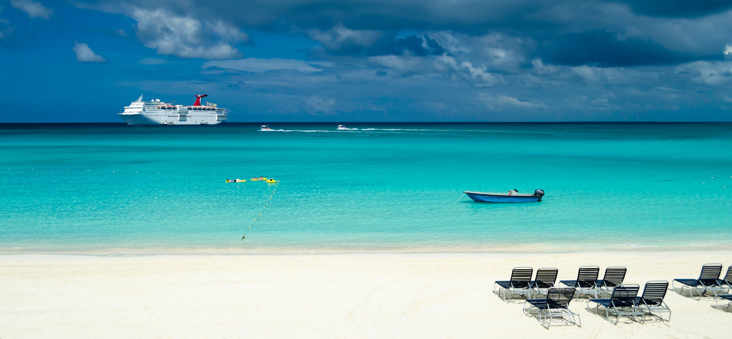 A white sandy beach with lounge chairs overlooking a turquoise bay, a small boat, and a cruise ship in the distance.