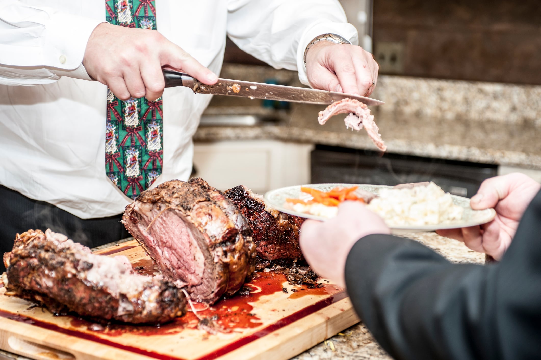 A person in a white dress shirt and patterned tie carves a slice of roast beef and places it onto a plate held by another person.