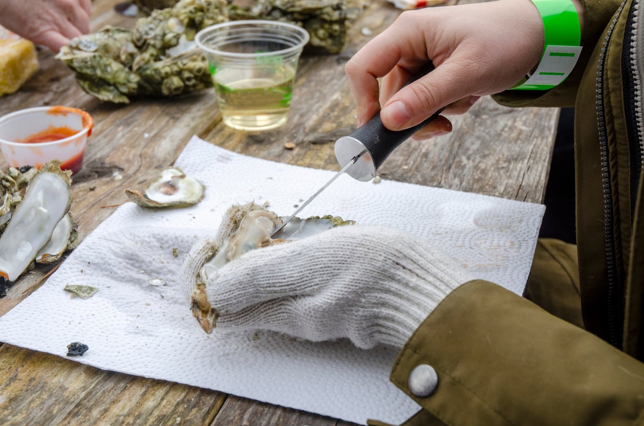 A person wearing a glove shucks an oyster with a small knife on a wooden table.