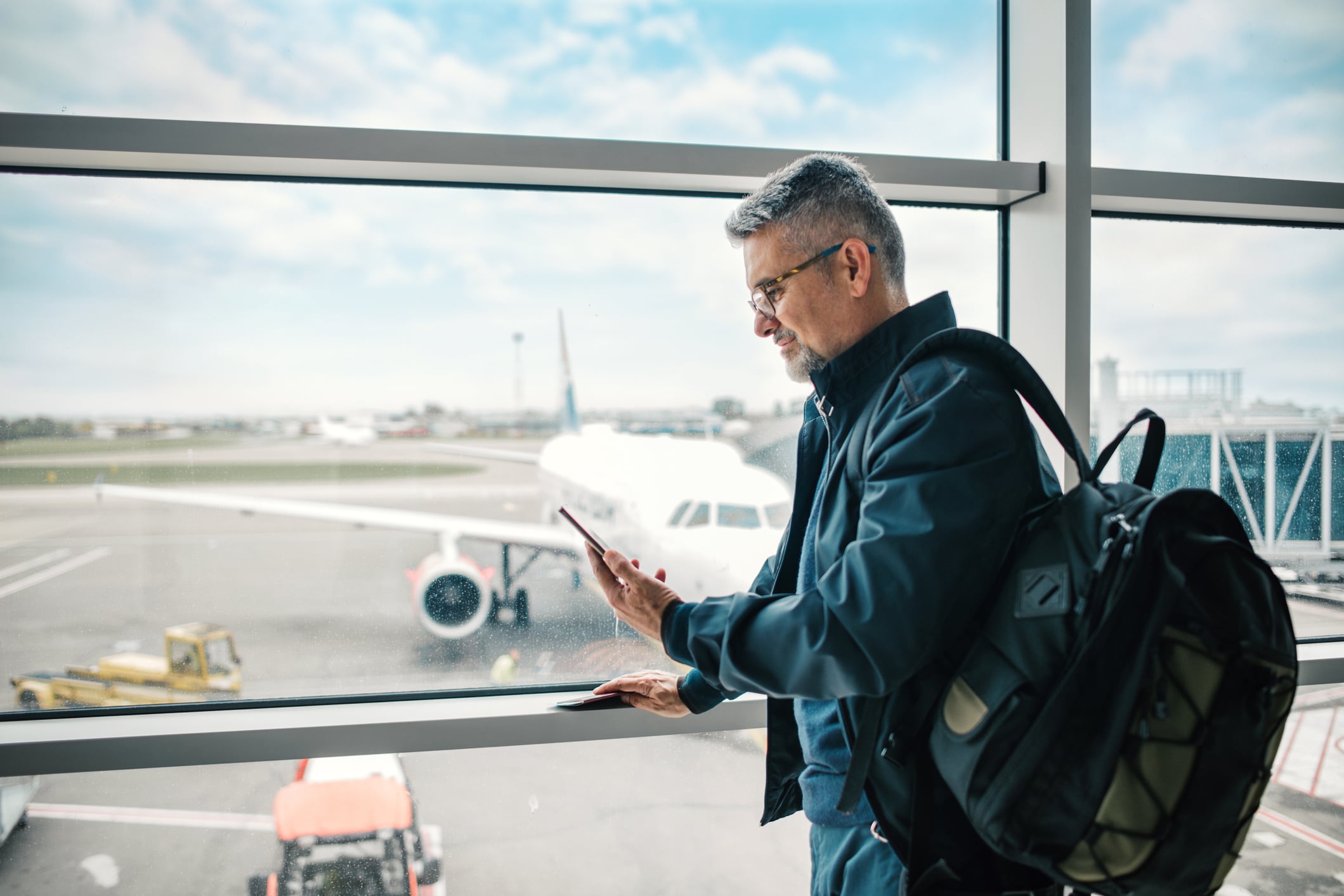 A man with a backpack stands by an airport window looking at his smartphone, with a parked airplane visible on the tarmac outside.