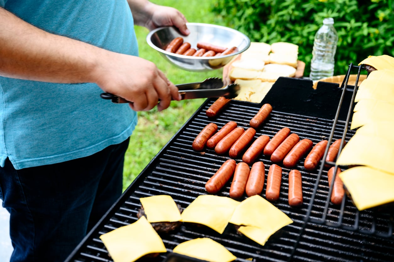 A person uses tongs to place hot dogs onto a grill filled with hot dogs and cheeseburgers.