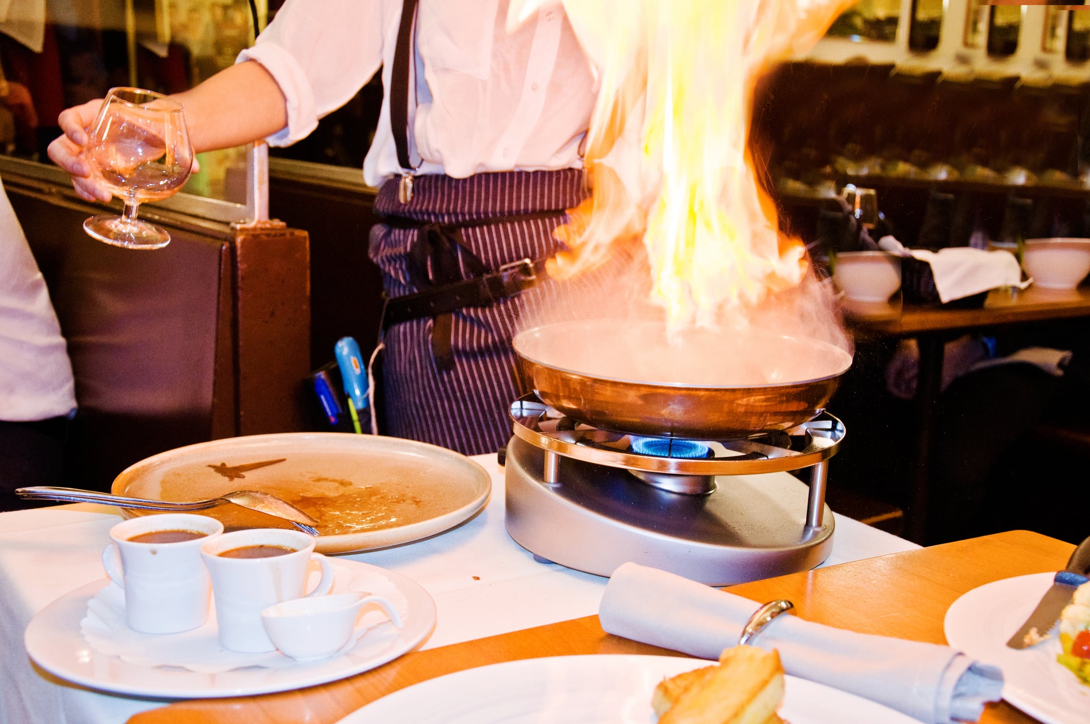 A server in a tableside dining bistro apron flambés a dish in a copper pan on a tabletop burner, while holding a glass of amber liquid.