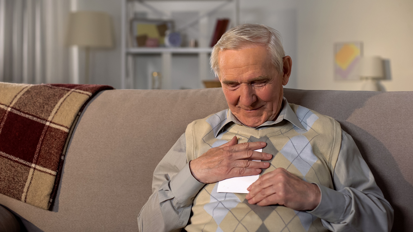 An elderly man sitting on a sofa, smiling while holding a letter against his chest.