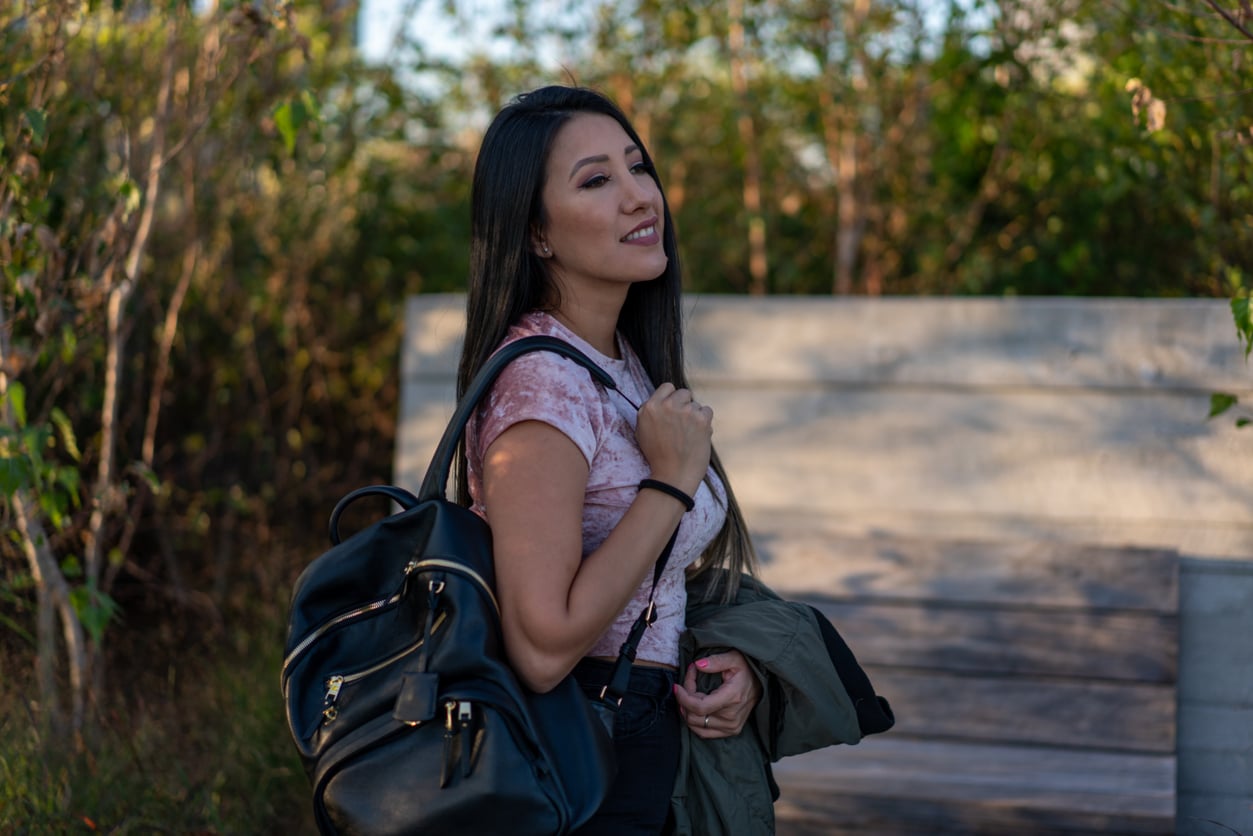 A smiling woman with long dark hair stands outdoors carrying a black leather backpack and holding a jacket over her arm.