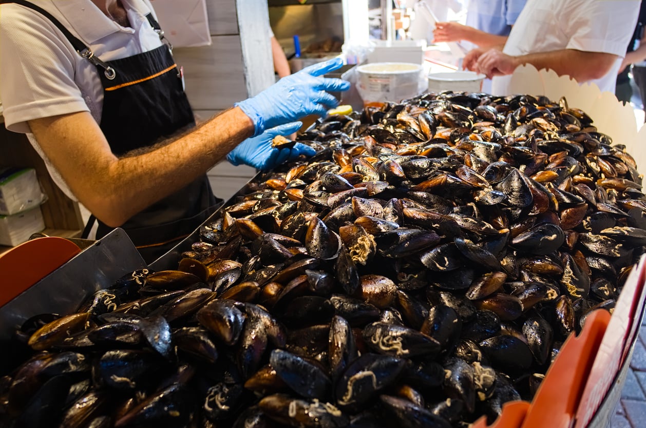 A food vendor wearing blue gloves scoops a large pile of cooked mussels from a metal tray.