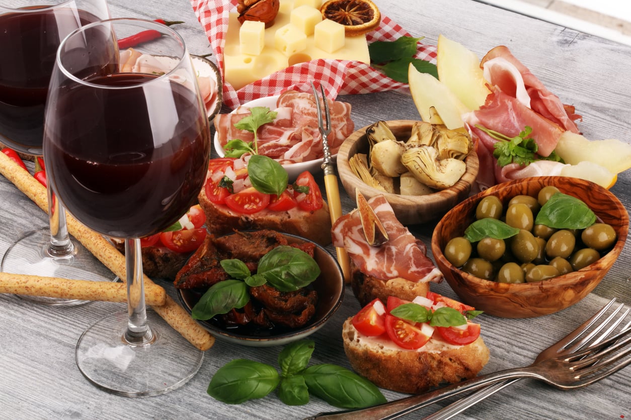 Two glasses of red wine accompanied by a charcuterie board featuring bruschetta, olives, artichoke hearts, prosciutto, cheese, and breadsticks.
