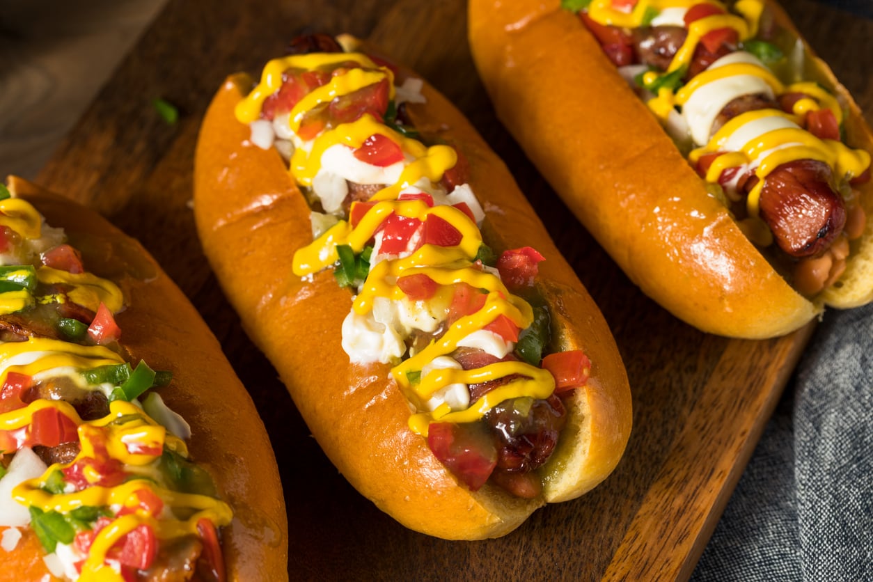 Three hot dogs in buns topped with yellow mustard, chopped tomatoes, green peppers, and sour cream on a wooden board.
