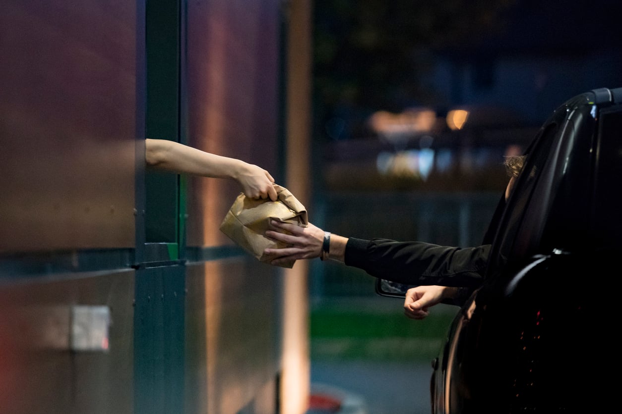 A drive-thru window worker hands a paper bag to a person waiting in a car.