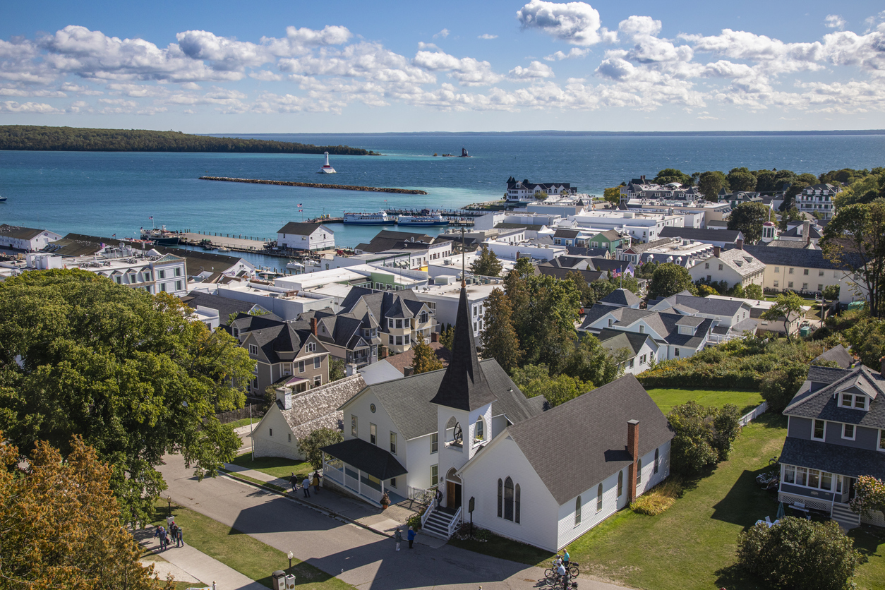 Aerial view of a coastal town with a church in the foreground, white buildings, a harbor, docks, and a lighthouse along the blue shoreline under a partly cloudy sky, surrounded by green trees and distant land.