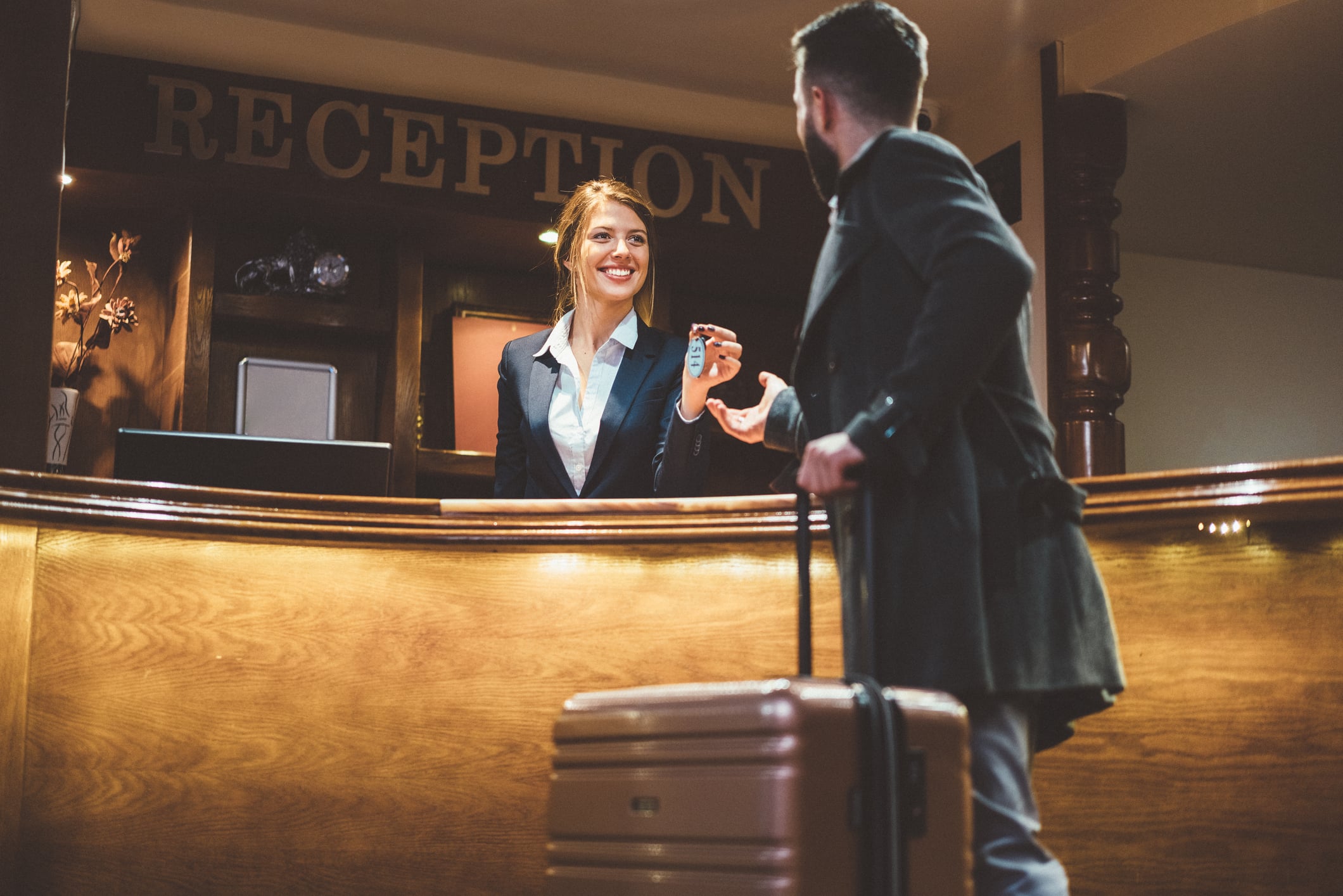 A smiling hotel receptionist hands a room key to a guest standing at the front desk with a suitcase.