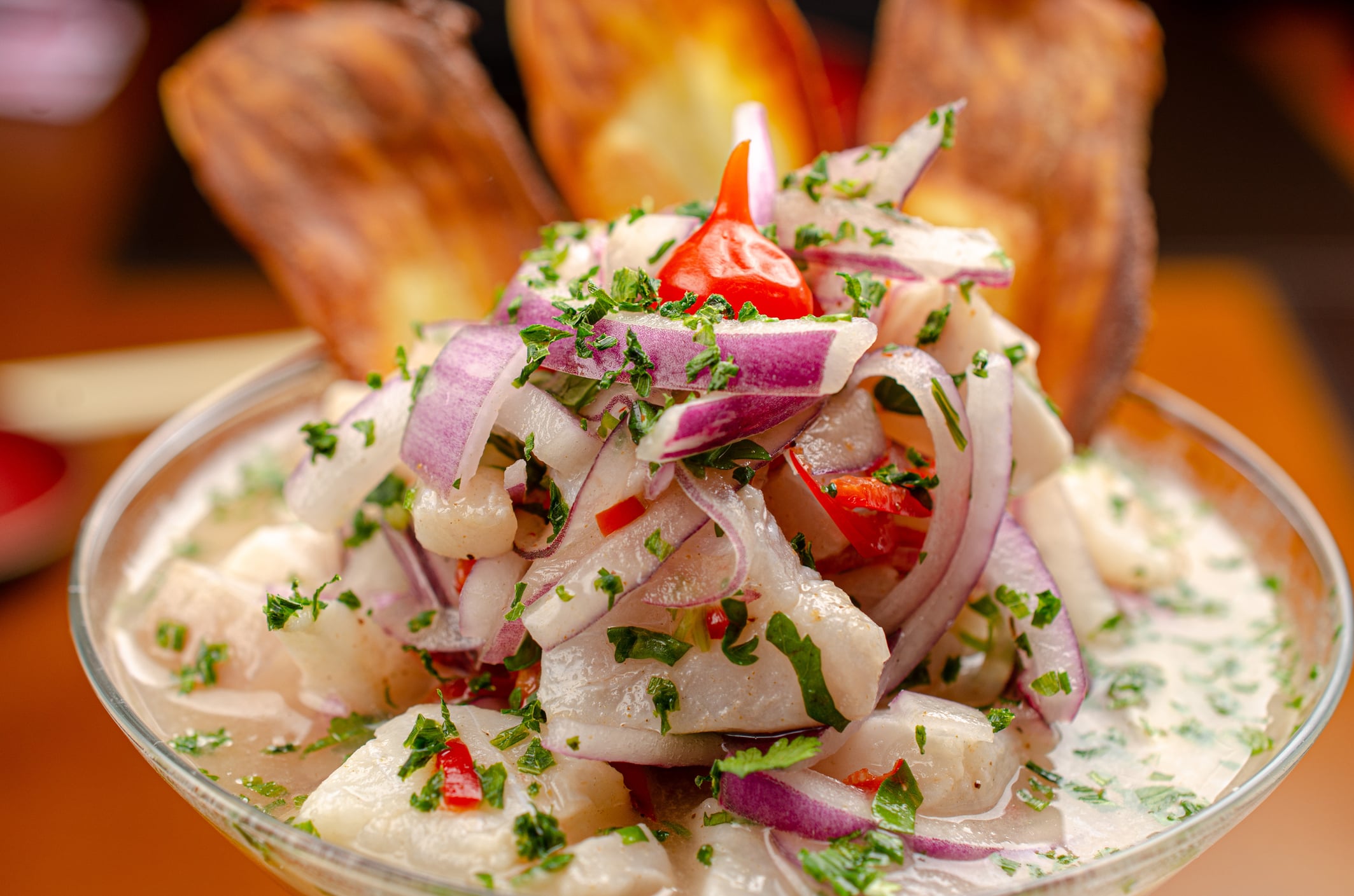 A glass bowl filled with ceviche topped with red onion slices, herbs, and a single red chili pepper, served with fried plantain chips.