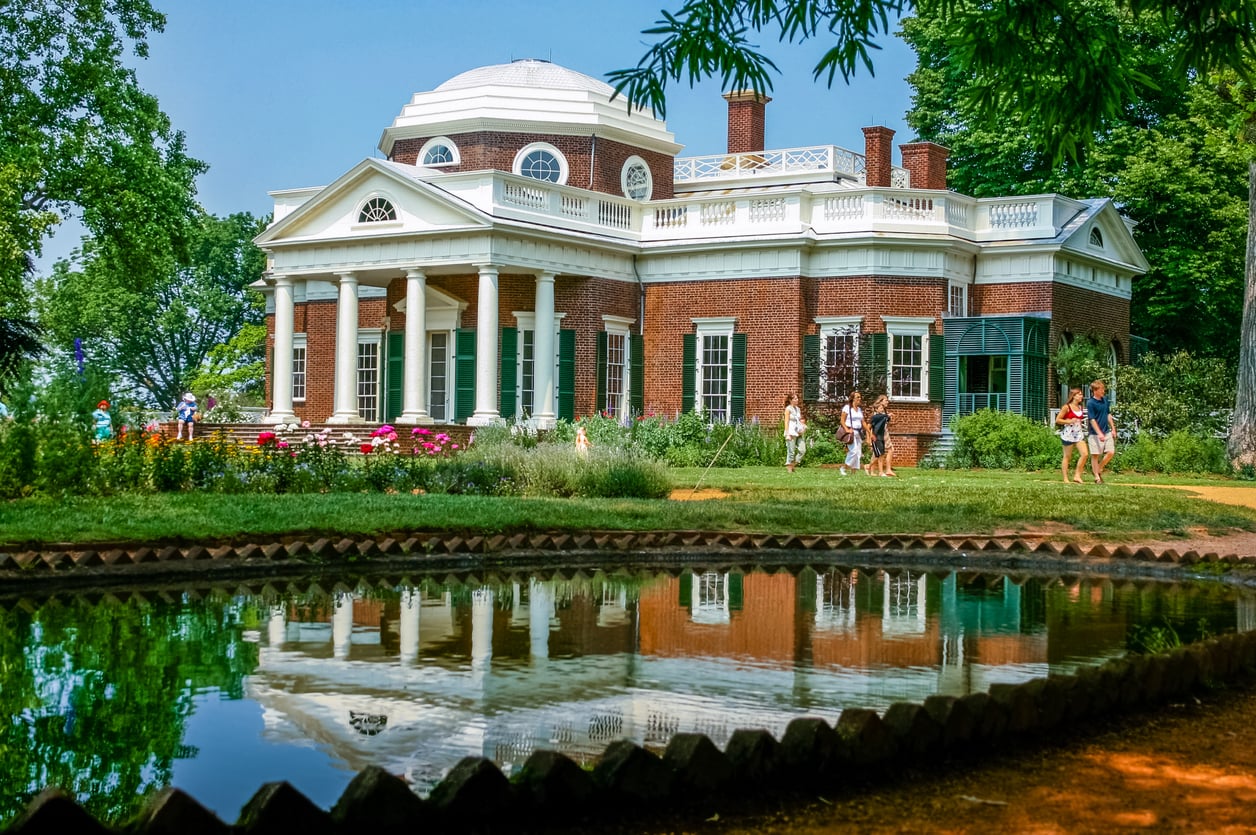 Monticello, a historic brick mansion with a white dome and columns, viewed across a reflecting pool with visitors walking on the lawn.