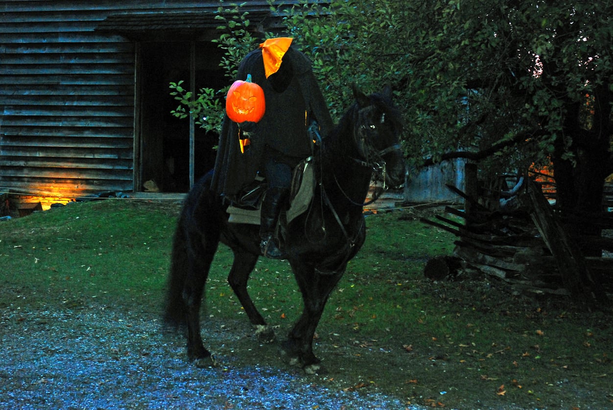 A headless horseman in a black cloak sits on a black horse, holding a glowing orange jack-o’-lantern.